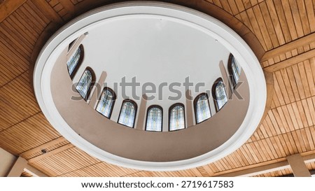 Low-angle view of a circular architectural dome with stained glass windows set into a wooden plank ceiling. Modern interior design with elegant lighting and symmetry. High quality.