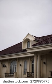 Low-angle view of a classical building featuring a red tiled roof, arched dormer window, and elegant architectural details against an overcast sky, highlighting timeless design and formal architecture