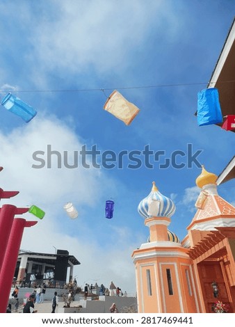 Low-angle view of colorful hanging lanterns strung across a clear blue sky, featuring whimsical onion-dome architecture and torii gate structures at a theme park.