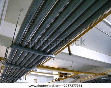 A low-angle view of a complex industrial ceiling infrastructure features multiple parallel dark gray cylindrical conduits mounted securely to a white concrete support beam using a metal rack system. The pipes feature faint printed Japanese characters and technical markings, including text that translates to '80mm', indicating size specifications for the utility lines. Below the primary conduit bank, a network of thinner, bright yellow pipes runs across the ceiling, possibly representing a fire sprinkler system or gas supply lines. The stark contrast between the muted, industrial gray pipes and the vivid yellow accents, set against a sterile, clean white ceiling, creates a functional and utilitarian aesthetic. This scene captures the hidden mechanical veins of a modern commercial or institutional building, emphasizing themes of engineering, maintenance, and urban infrastructure.