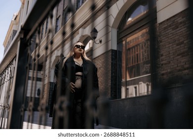 Low-angle view of confident plus size woman in black erotic lingerie and leather trench coat. Stylish curvy model on urban street near building and decorative railing, promoting body positivity.