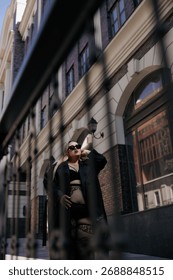Low-angle view of confident plus size woman in black erotic lingerie and leather trench coat. Stylish curvy model on urban street near building and decorative railing, promoting body positivity.