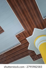 Low-angle view of a decorative building ceiling featuring a central yellow pillar surrounded by symmetrical geometric patterns and wood-textured ceiling panels. The architectural composition highlights interior design aesthetics, structural elements, symmetry, and modern construction concepts. Ideal for themes related to architecture, building interiors, construction, engineering, public facilities, and decorative ceiling design.