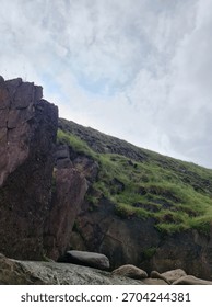 Low-angle view of a dramatic coastal scene with massive dark rocks, a grassy steep slope, and boulders on the shore. Highlights the rugged geology and textures of the cliff face.