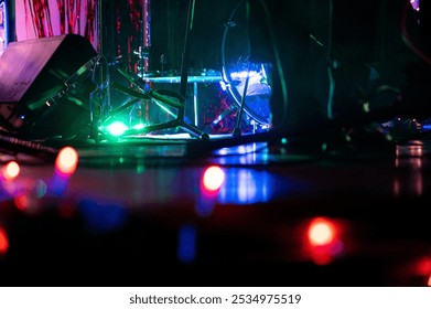 Low-angle view of a drum set and mic stand on a dimly lit stage with colorful Christmas lights and dramatic lighting. A raw, intimate house show vibe with cables, glow, and gritty energy.