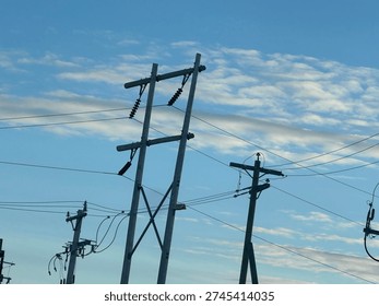 Low-angle view of electrical substation frames, transmission lines, and utility poles silhouetted against a textured evening sky. Industrial energy infrastructure contrasts with layered clouds, highlighting grid systems, power distribution, and modern utility networks.