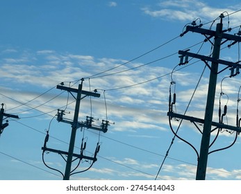 Low-angle view of electrical substation frames, transmission lines, and utility poles silhouetted against a textured evening sky. Industrial energy infrastructure contrasts with layered clouds, highlighting grid systems, power distribution, and modern utility networks.