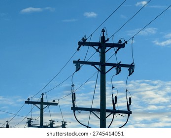 Low-angle view of electrical substation frames, transmission lines, and utility poles silhouetted against a textured evening sky. Industrial energy infrastructure contrasts with layered clouds, highlighting grid systems, power distribution, and modern utility networks.
