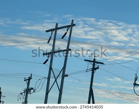 Low-angle view of electrical substation frames, transmission lines, and utility poles silhouetted against a textured evening sky. Industrial energy infrastructure contrasts with layered clouds, highlighting grid systems, power distribution, and modern utility networks.