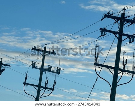 Low-angle view of electrical substation frames, transmission lines, and utility poles silhouetted against a textured evening sky. Industrial energy infrastructure contrasts with layered clouds, highlighting grid systems, power distribution, and modern utility networks.
