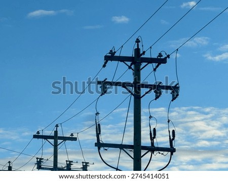 Low-angle view of electrical substation frames, transmission lines, and utility poles silhouetted against a textured evening sky. Industrial energy infrastructure contrasts with layered clouds, highlighting grid systems, power distribution, and modern utility networks.