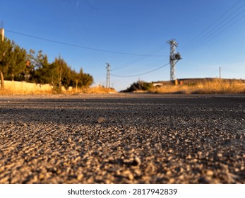 Low-angle view of an empty asphalt road stretching through a rural landscape with trees, utility poles, and a clear blue sky during golden hour.