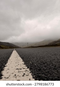 Low-angle view of an empty road with a strong leading line perspective, stretching through a misty mountain landscape under dramatic cloudy skies. The textured asphalt and central road marking create a powerful sense of direction, journey, and movement. The atmospheric fog and surrounding hills enhance the mood of isolation and exploration, making it ideal for themes of travel, freedom, business concepts, and future direction.