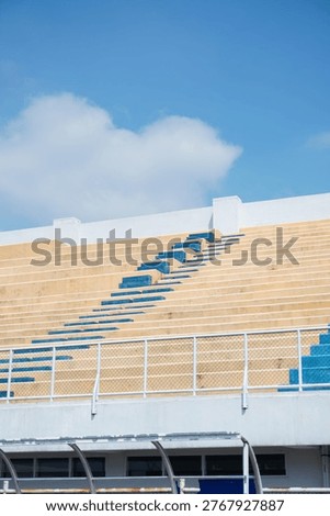 A low-angle view of empty stadium bleachers featuring rows of tan concrete steps with a central section of blue seats, set against a bright blue sky.
