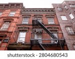A low-angle view of a facade of a historic brick tenement apartment building in New Your City, Brooklyn, with  a metal zig-zag fire escape running down the center. An old multi-story red brick residential building in NYC.