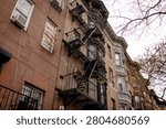 A low-angle view of a facade of a historic brick tenement apartment building in New Your City, Brooklyn, with  a metal zig-zag fire escape running down the center. An old multi-story red brick residential building in NYC.