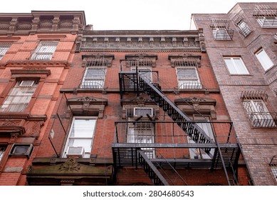 A low-angle view of a facade of a historic brick tenement apartment building in New Your City, Brooklyn, with  a metal zig-zag fire escape running down the center. An old multi-story red brick residential building in NYC.