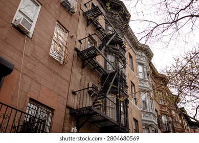 A low-angle view of a facade of a historic brick tenement apartment building in New Your City, Brooklyn, with  a metal zig-zag fire escape running down the center. An old multi-story red brick residential building in NYC.