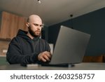 Low-angle view of focused programmer wearing dark hoodie typing code on laptop computer, sitting at table near bright window in contemporary urban apartment with minimalist design and soft daylight.
