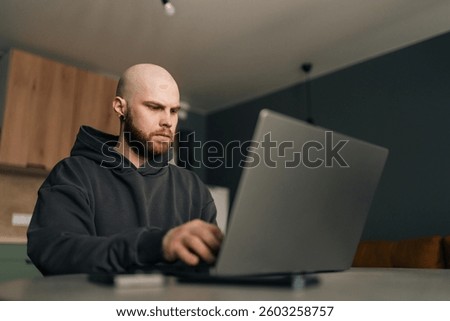 Low-angle view of focused programmer wearing dark hoodie typing code on laptop computer, sitting at table near bright window in contemporary urban apartment with minimalist design and soft daylight.