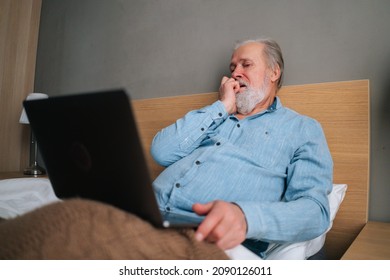 Low-angle View Of Frustrated Gray-haired Senior Male With Beard Using Laptop Computer Sitting On Bed At Home. Handsome Bearded Mature Older Man In Pajamas Having Online Video Call Via Notebook.