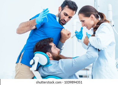 Low-angle View Of A Funny Dentist Or Dental Surgeon Acting Crazy In Front Of His Female Assistant, While Having A Patient On The Chair In The Dental Office