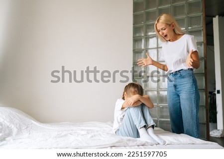 Low-angle view of furious blonde mom scolding, raising voice, yelling for bad behavior to sad crying little girl covering face with palm sitting on bed at home. Concept of mother daughter problem.
