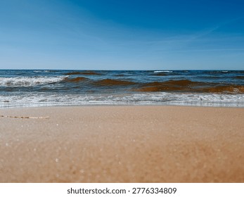 Low-angle view of gentle ocean waves rolling onto a sandy beach beneath a clear blue sky. Clean horizon and natural light create a calm, minimal seascape ideal for travel, relaxation, and summer background use.