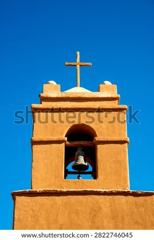 Low-angle view of the iconic adobe bell tower topped with a simple wooden cross at the Church of San Pedro de Atacama, Chile, set against a vibrant clear blue sky. Historic colonial architecture.