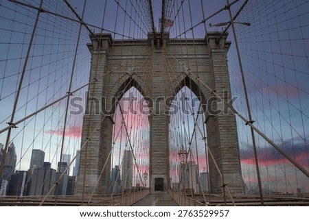 Low-angle view of iconic Brooklyn Bridge with stone towers, suspension cables and American flag against dramatic pink and blue sunset sky with Manhattan skyline in background.