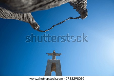 Low-angle view of the iconic Christ statue at the Sanctuary of Christ the King in Lisbon, Portugal. Dramatic perspective with clear blue sky and foreground chain detail, capturing the monument’s grandeur and spiritual symbolism.