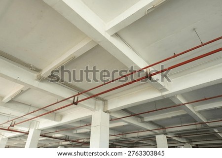 A low-angle view of an industrial building ceiling featuring a grid of white concrete beams, exposed red fire sprinkler piping, and electrical conduits.
