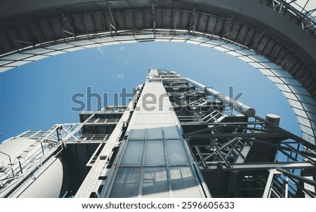 A low-angle view of an industrial launch pad with metal structures, pipes, and a blue sky background. The composition emphasizes height, engineering, and technology