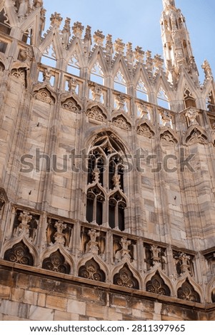 A low-angle view of the intricate Gothic marble architecture, spires, and traceried windows of the Milan Cathedral under a clear blue sky.