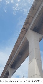 A low-angle view of a large concrete overpass against a bright blue sky. The structure’s pillars and underside highlight modern engineering, urban infrastructure, and industrial architecture