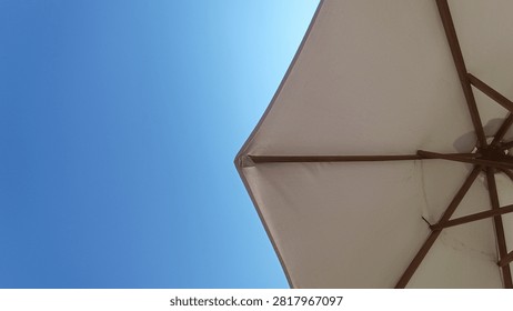 A low-angle view of a light-colored beach umbrella providing shade against a brilliant clear blue sky on a sunny day, highlighting its structural ribs.