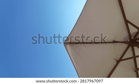 A low-angle view of a light-colored beach umbrella providing shade against a brilliant clear blue sky on a sunny day, highlighting its structural ribs.