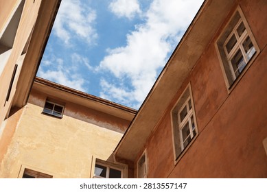 A low-angle view looking up at the corners of two old buildings with symmetrical windows, framing a bright blue sky with scattered clouds. The strong geometric lines create a bold architectural composition.