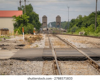 A low-angle view looking down a straight railway track leading past a gravel crossing towards distant concrete coal towers under a cloudy sky