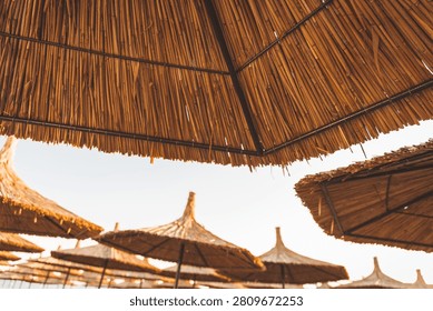 A low-angle view looking out from underneath a textured straw beach umbrella at a seaside resort. The warm golden hour light creates a relaxing summer vacation atmosphere against a pale evening sky