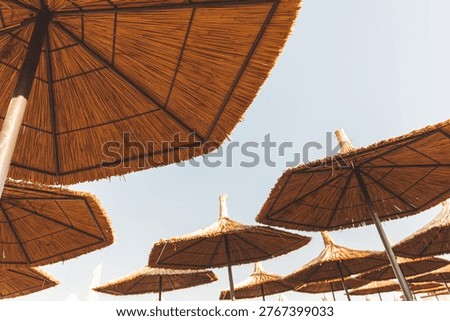 A low-angle view looking out from underneath a textured straw beach umbrella at a seaside resort. The warm golden hour light creates a relaxing summer vacation atmosphere against a pale evening sky