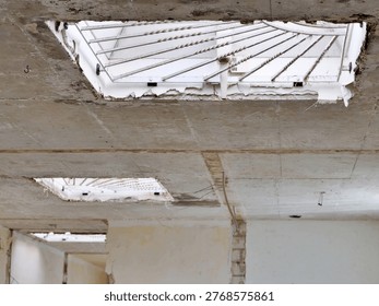 Low-angle view looking up at a rectangular opening in a weathered concrete
ceiling or wall. The opening is protected by a decorative white metal grate
featuring a semi-circular sunburst design with radiating rays. The surrounding
architectural structure shows raw textures, age, and slight decay. Behind the
security screen, a bright, neutral surface is visible, creating a geometric
contrast.