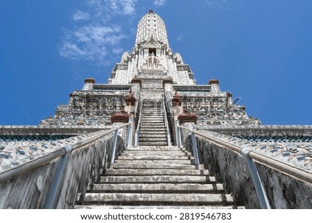 Low-angle view looking up the steep stairs of the ornate white prang at Wat Arun temple against a blue sky in Bangkok, Thailand. Iconic Buddhist architecture landmark.