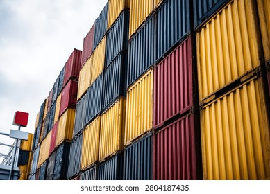 A low-angle view of a massive stack of shipping containers in various colors, including yellow, red, and blue, at a port.