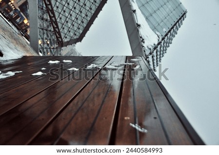 a low-angle view of a modern architectural structure with glass panels reflecting the overcast sky, and a wooden bench in the foreground with patches of snow on it