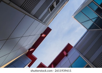 Low-angle view of modern architecture with sharp angles, red accents, and glass windows against a bright, slightly cloudy sky.