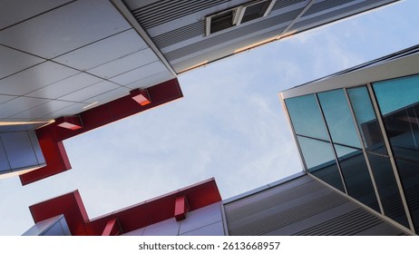 Low-angle view of modern architecture with sharp angles, red accents, and glass windows against a bright, slightly cloudy sky.