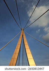 A low-angle view of a modern cable-stayed bridge against a blue sky. The cables and structure form a symmetrical pattern. Captured in Thailand on June 16, 2025.