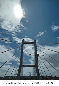 Low-angle view of a modern cable-stayed bridge against a dramatic blue sky with bright sunlight and scattered clouds. Urban infrastructure, transportation, and architectural engineering concept.