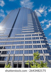 Low-angle view of a modern skyscraper with reflective glass panels against a bright blue sky and wispy clouds. Urban architecture and business concept in a metropolitan setting.
